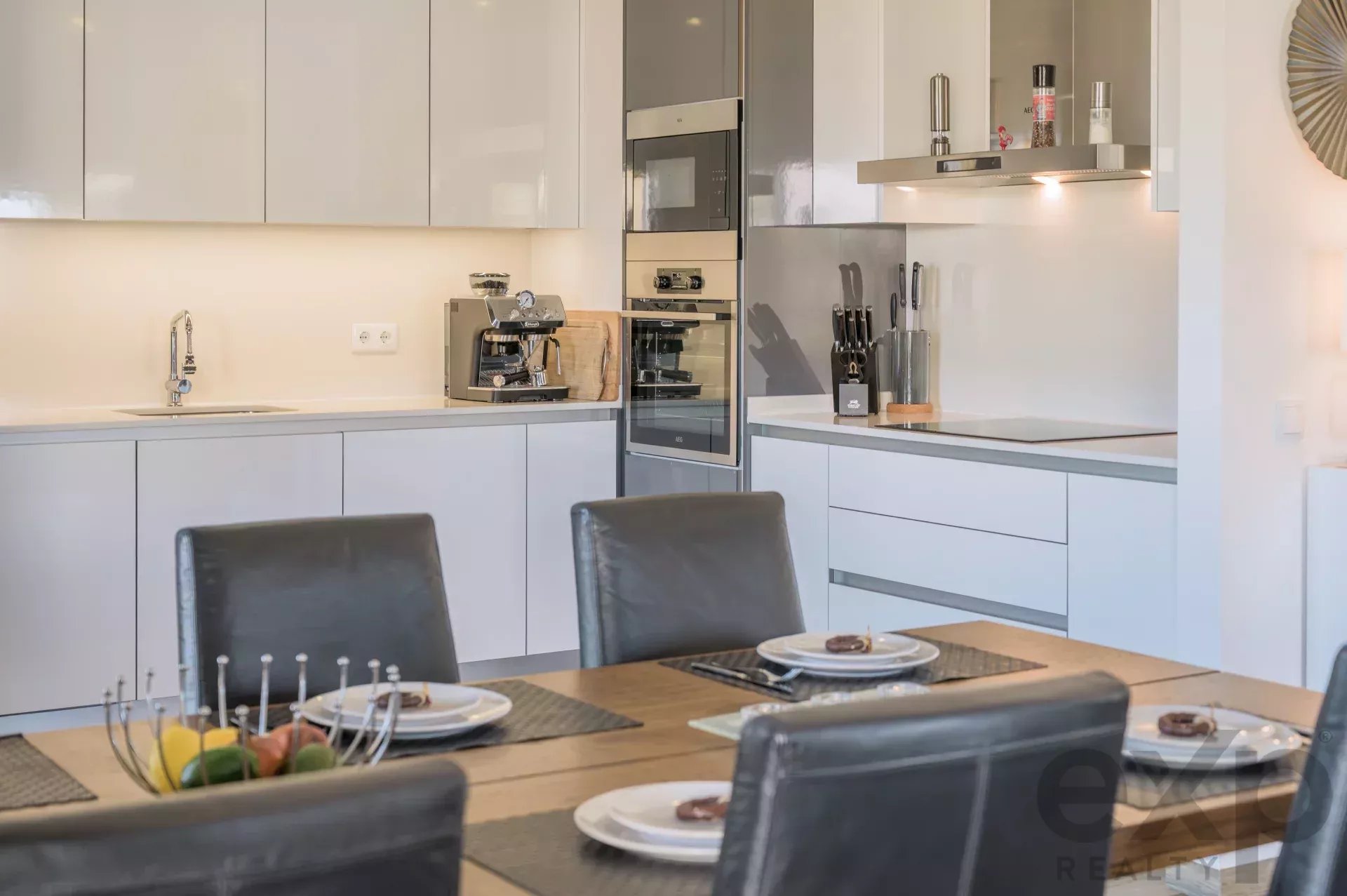 Modern kitchen with white glossy cabinets, built‑in oven and microwave, and a countertop coffee machine beside a knife block on the right.