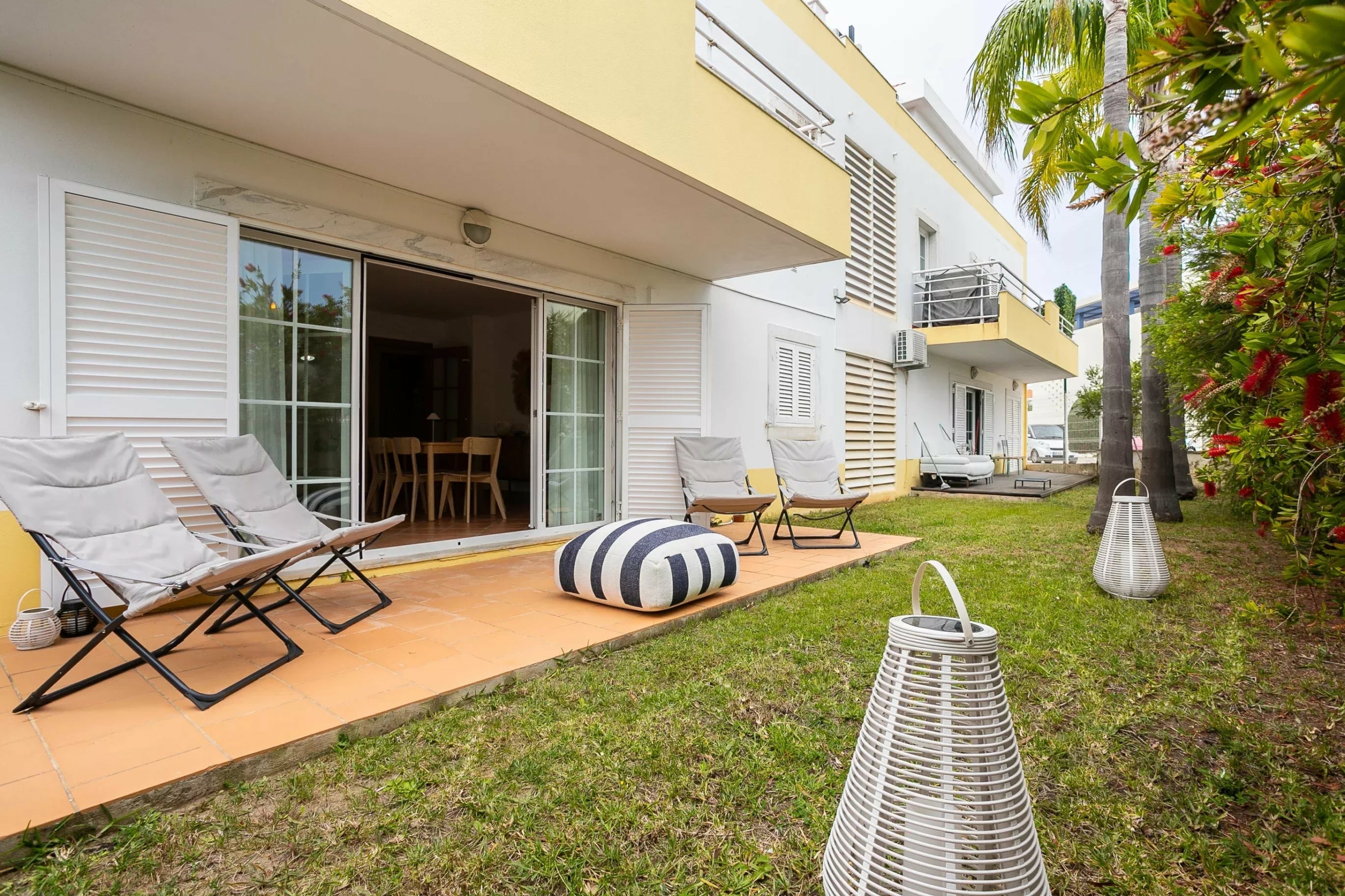 Outdoor patio with white lounge chairs on a tiled deck beside a pale-yellow apartment building and tropical plants on the right.