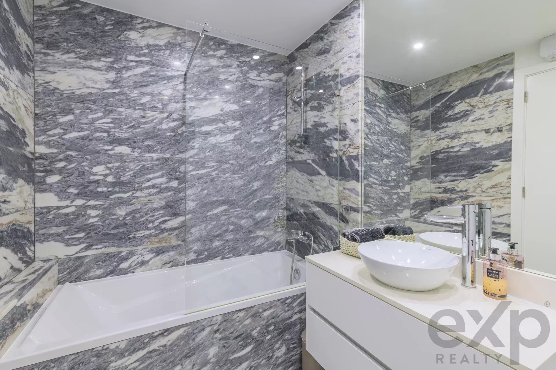 Luxurious marble-tiled bathroom with a glass shower enclosure, white bathtub, and dual white vessel sinks on a white vanity.