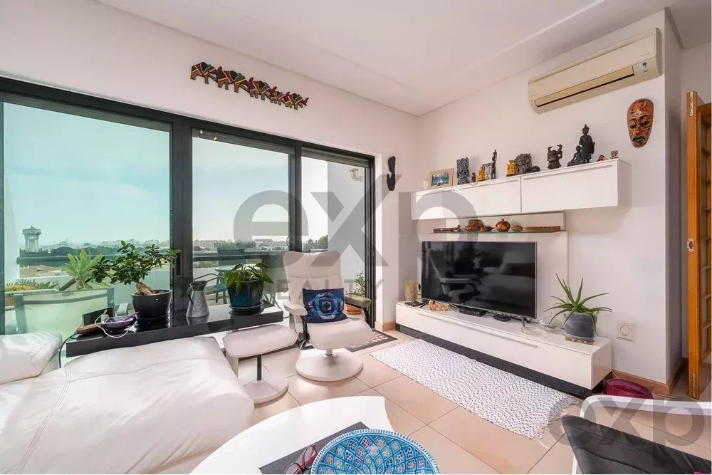 Bright living room with large glass doors to a balcony, white sofa, and a wall-mounted TV on a minimalist white entertainment unit with plants nearby.