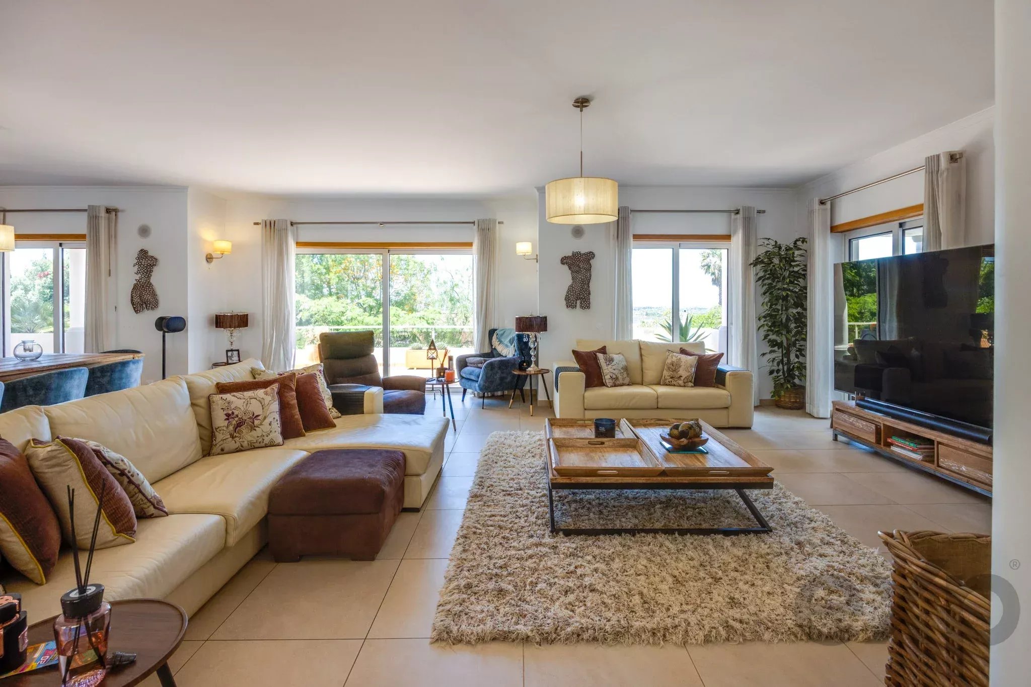 Spacious living room with cream sofas, brown cushions, a shaggy beige rug, and large windows with white curtains letting in sunlight.