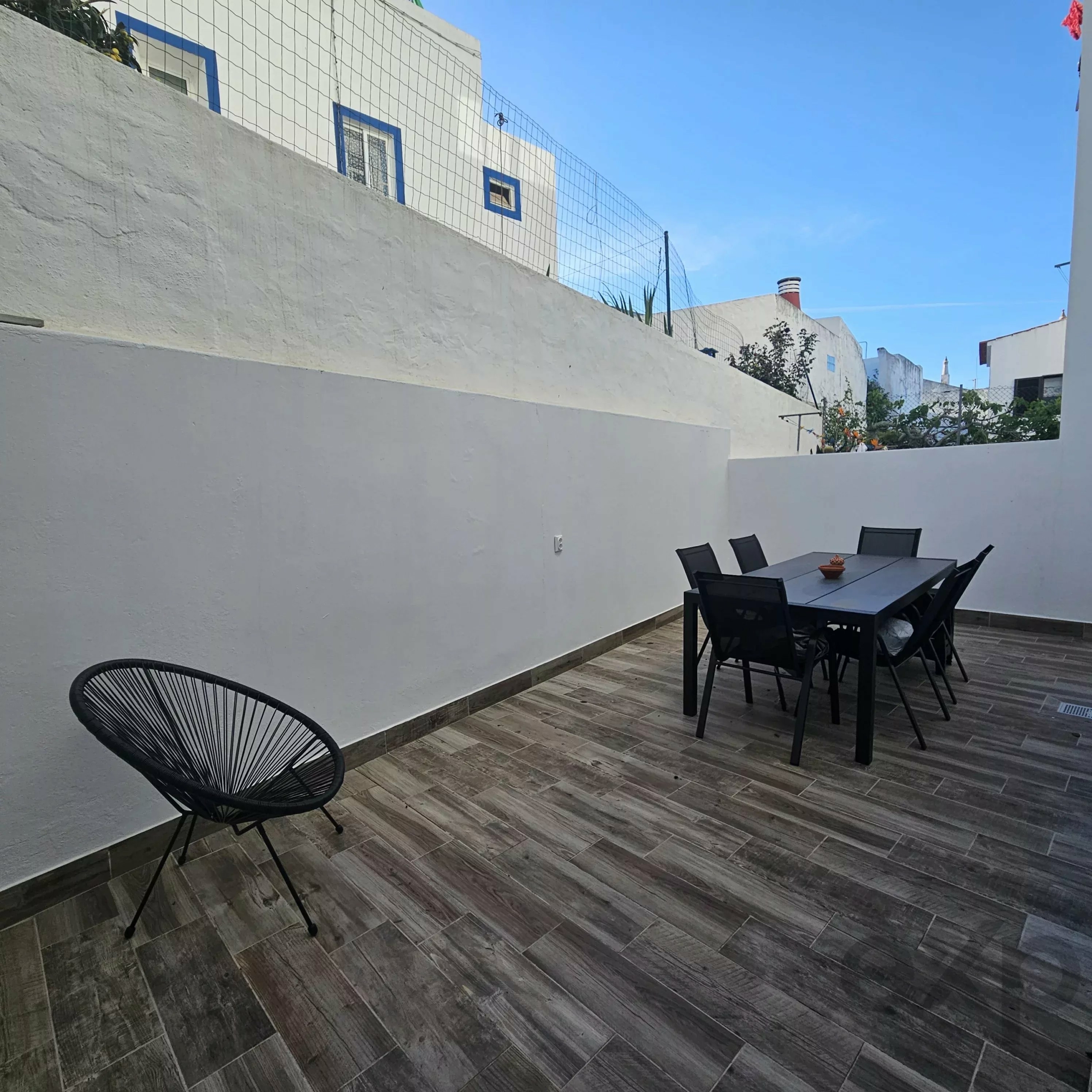 Outdoor dining patio with a black wire lounge chair in the foreground, a dark table and chairs on wood-like decking, white walls, and a clear blue sky.