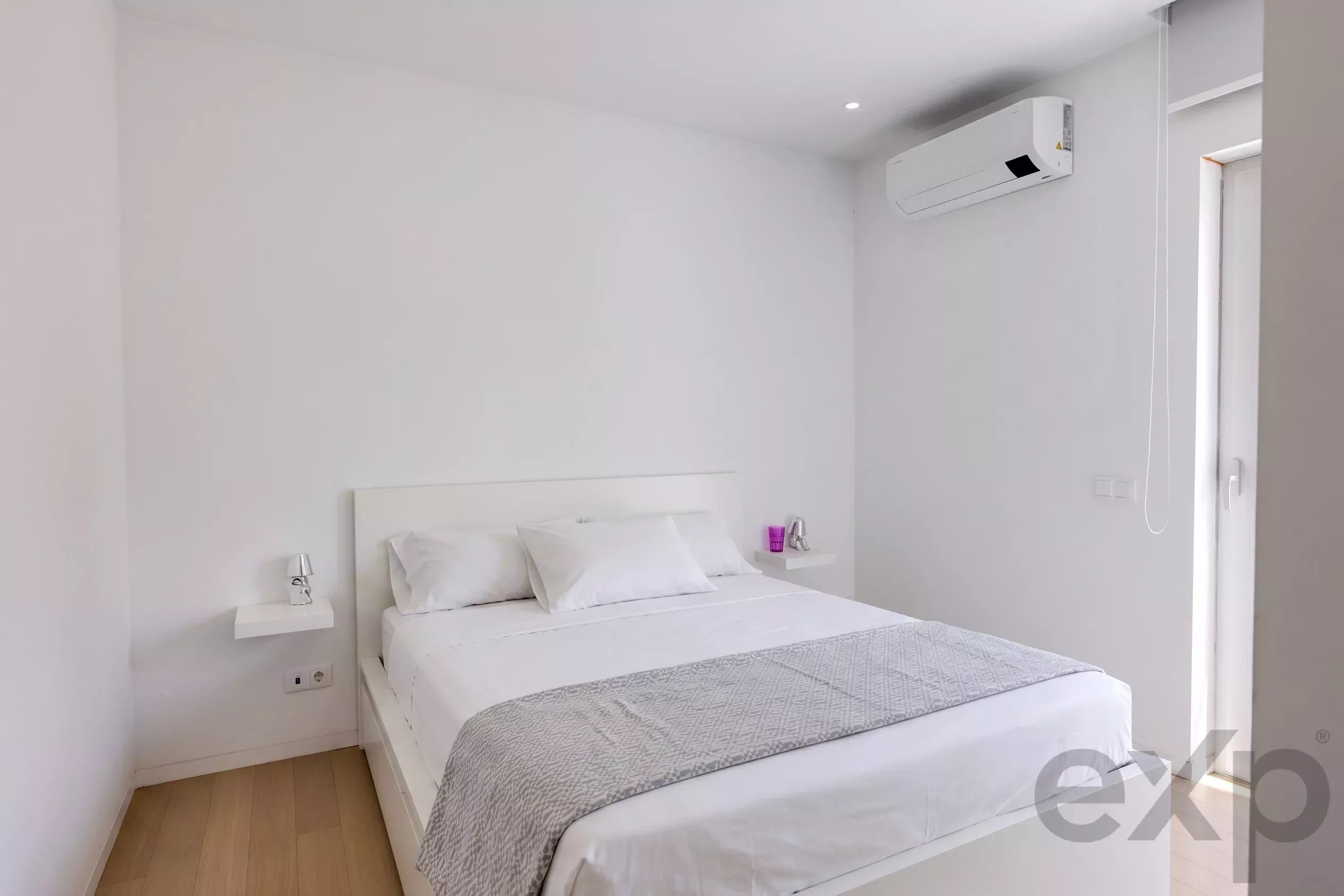 Modern white hotel bedroom with a bed, two floating nightstands, and a wall-mounted air conditioner.