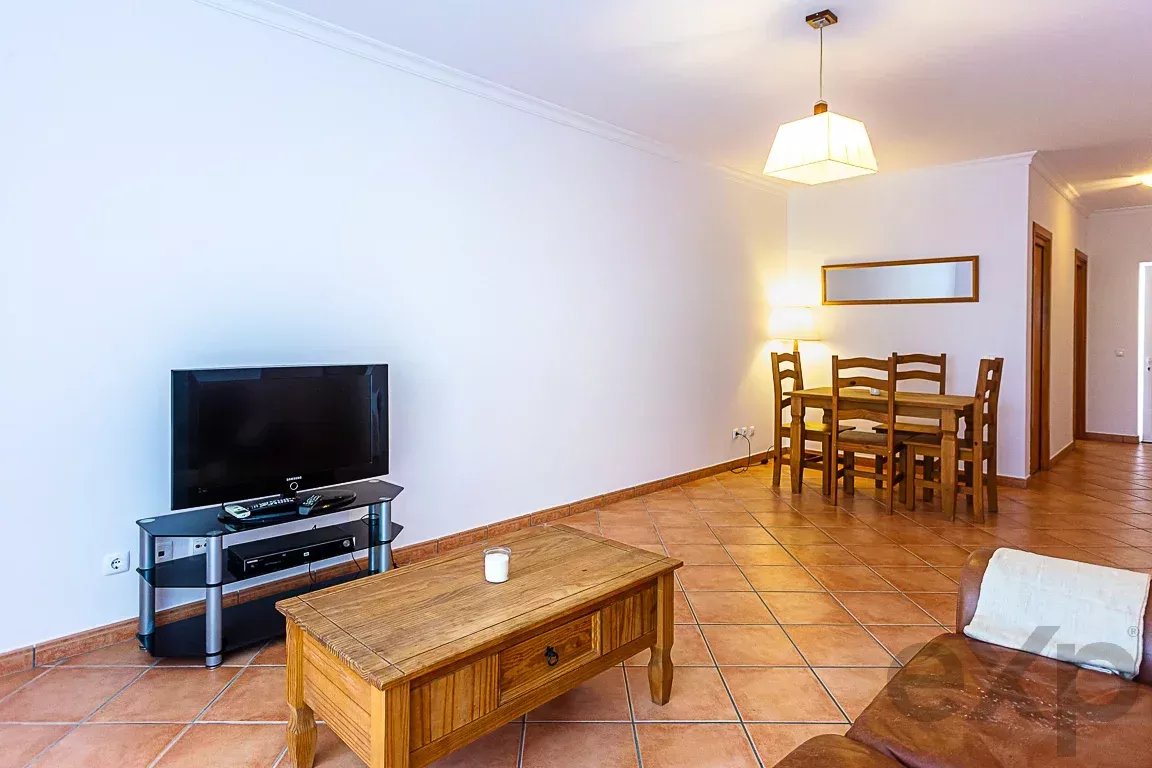 Open-plan living room with a wooden coffee table, sofa on the right, and TV on a glass stand; dining table and chairs visible in the background.
