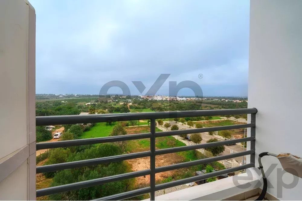 Balcony railing frames a rural landscape with green fields, trees, and a distant town under a cloudy sky.