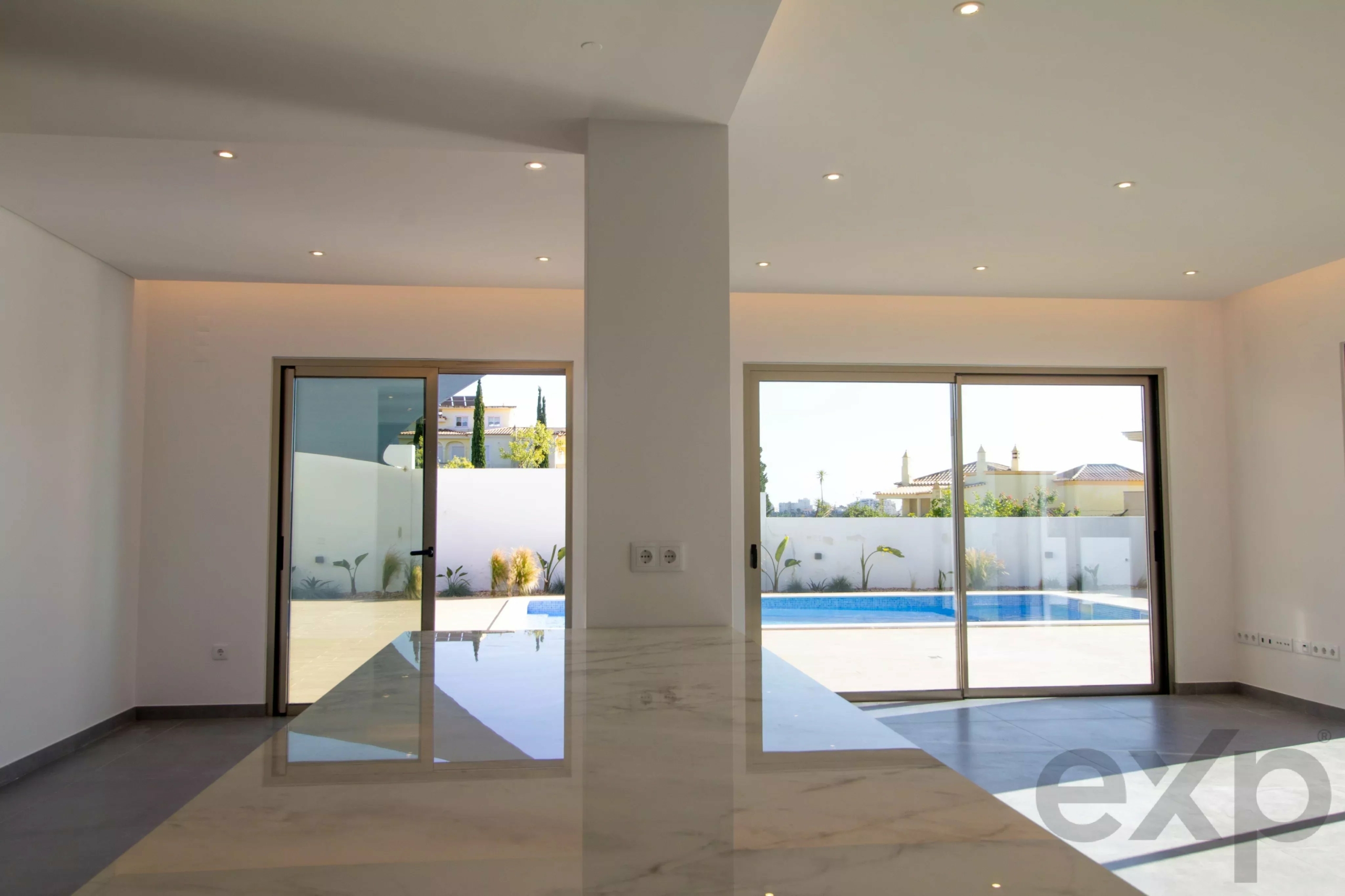 Bright open living area with a marble floor, white walls, a central supporting column, and large glass sliding doors opening to a sunny backyard with a pool and distant houses.