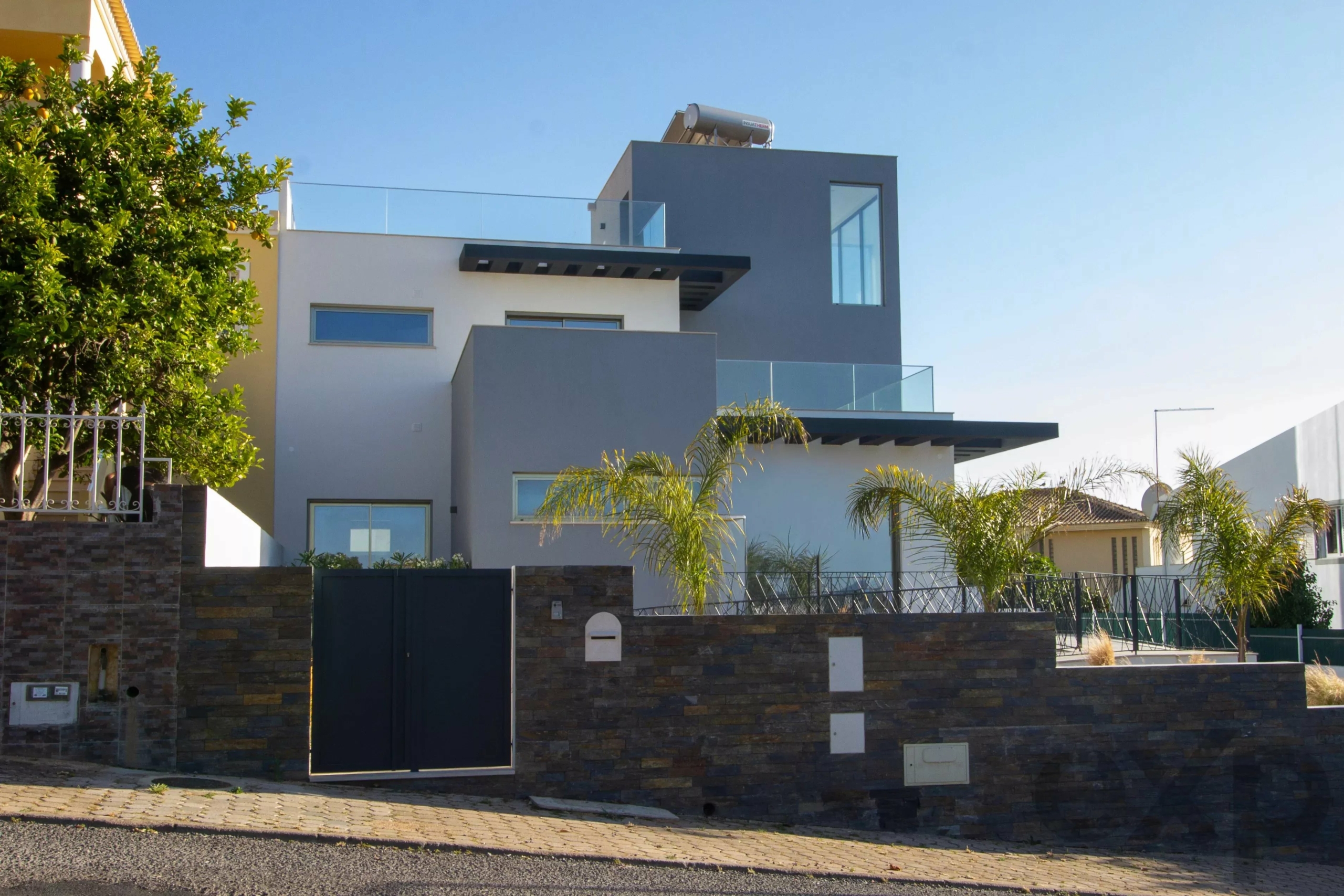 Modern multi‑story house with gray/white facade, glass balconies and palm trees in front, under a blue sky; contemporary design.