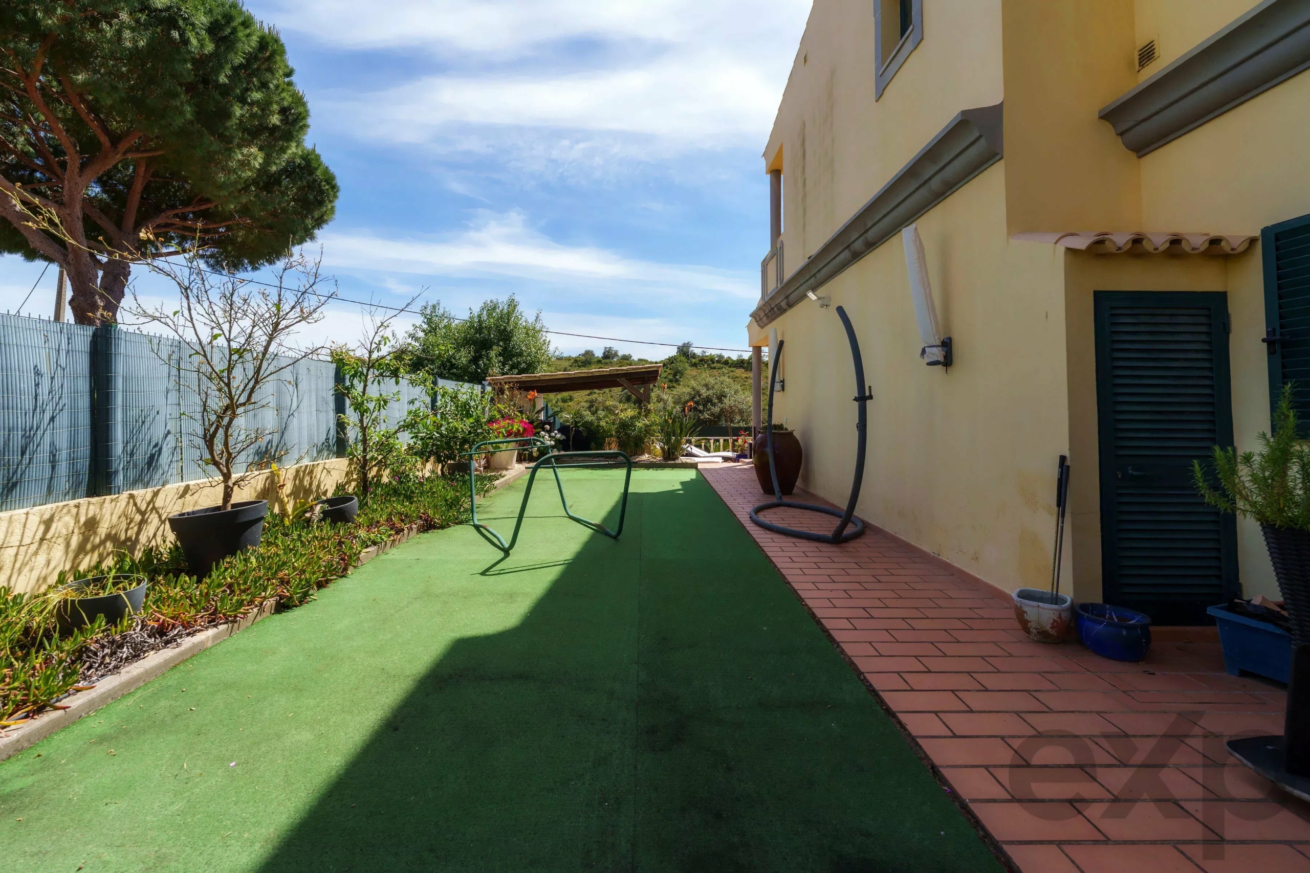 Backyard patio beside a yellow house, with green artificial turf, terracotta tiles, and potted plants under a blue sky.