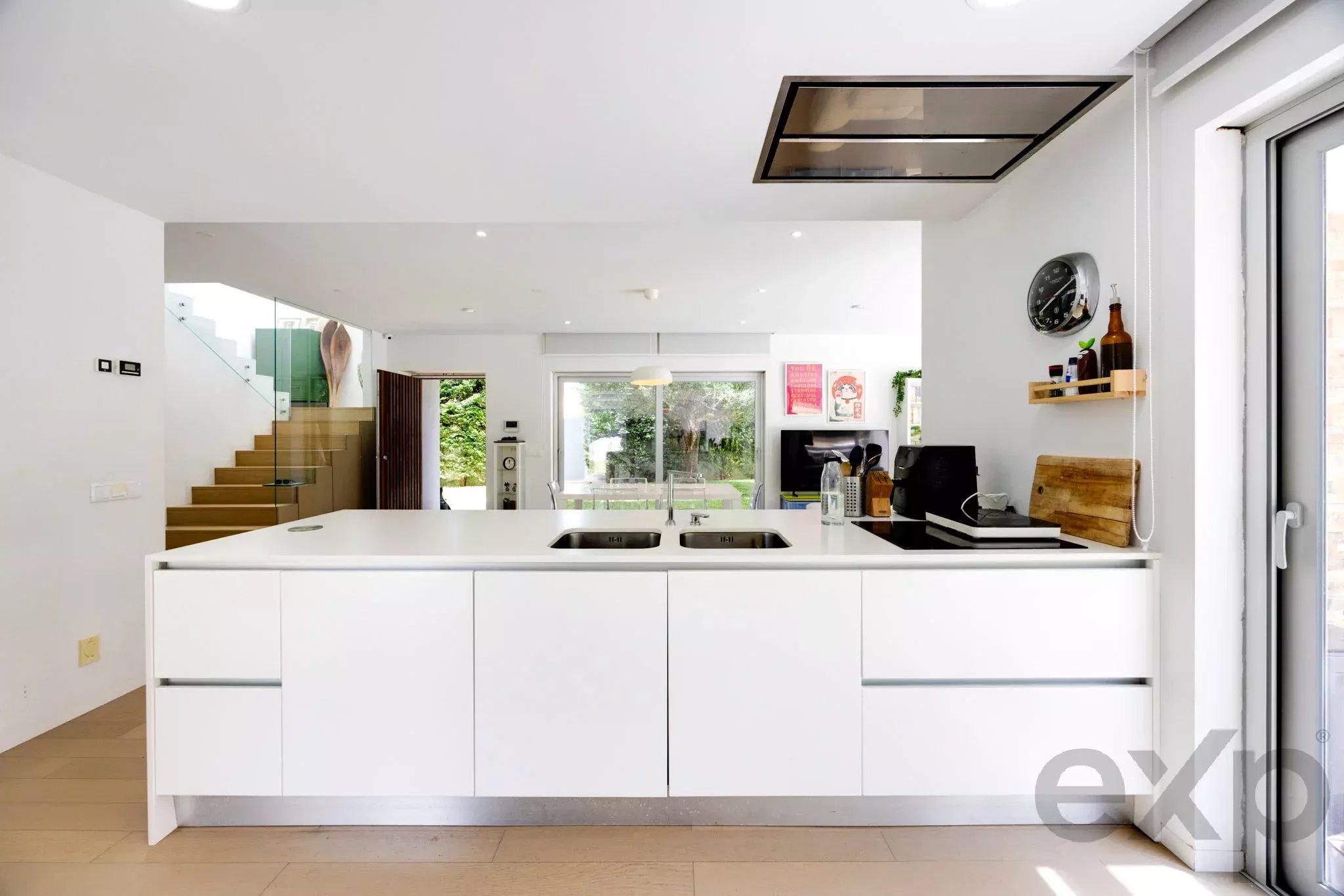Bright, minimalist kitchen with a white island sink, white cabinets, and stairs visible on the left in an open-plan space.