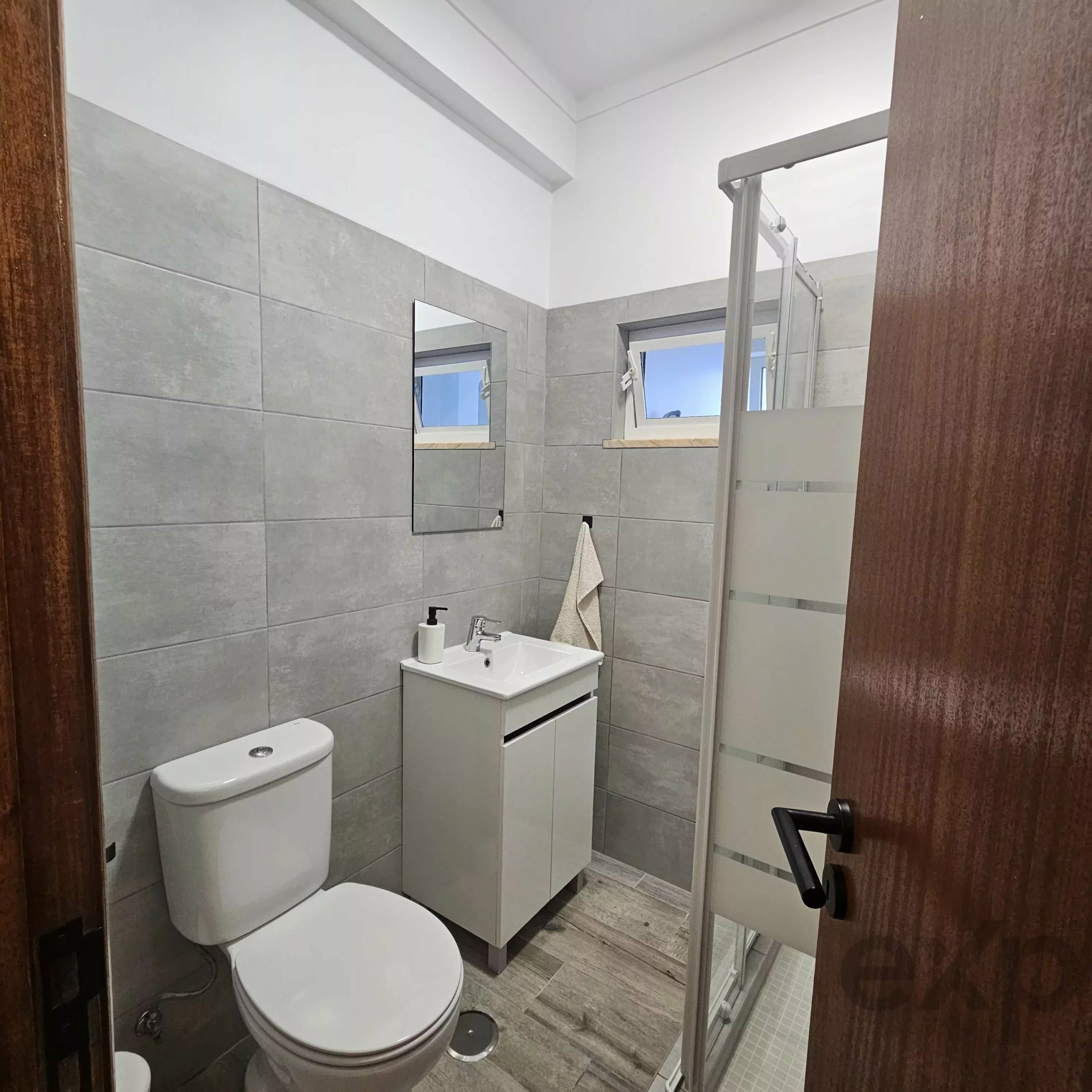 Small modern bathroom with gray tiled walls, white sink vanity, toilet, and a glass shower enclosure.