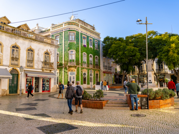 People walk in a sunny town square with a historic green tiled building and trees in the background.','A bustling pedestrian plaza with shops, a green tiled corner building, and seating areas.','Tourists and locals in a cobblestone square near a decorative building and a row of trees.','Public square scene with pedestrians, storefronts, and a large green-tiled building under a clear blue sky.