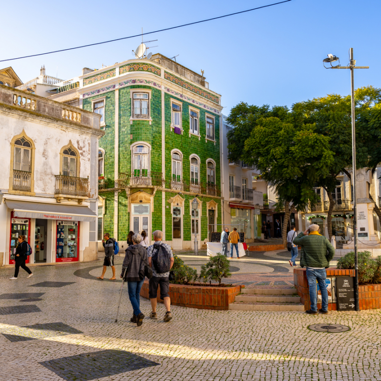 People walk in a sunny town square with a historic green tiled building and trees in the background.','A bustling pedestrian plaza with shops, a green tiled corner building, and seating areas.','Tourists and locals in a cobblestone square near a decorative building and a row of trees.','Public square scene with pedestrians, storefronts, and a large green-tiled building under a clear blue sky.