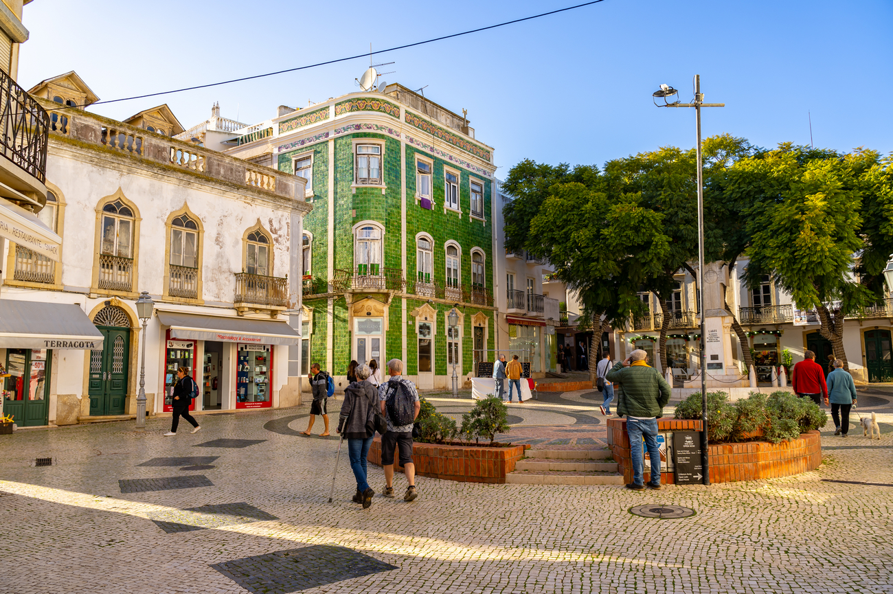 People walk in a sunny town square with a historic green tiled building and trees in the background.','A bustling pedestrian plaza with shops, a green tiled corner building, and seating areas.','Tourists and locals in a cobblestone square near a decorative building and a row of trees.','Public square scene with pedestrians, storefronts, and a large green-tiled building under a clear blue sky.