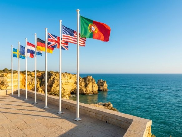 Row of international flags on poles along a sunny seaside promenade overlooking the ocean.