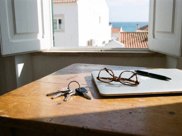 Keys, glasses and a laptop on a desk by an open shuttered window overlooking terracotta rooftops and the sea in Lagos, Algarve